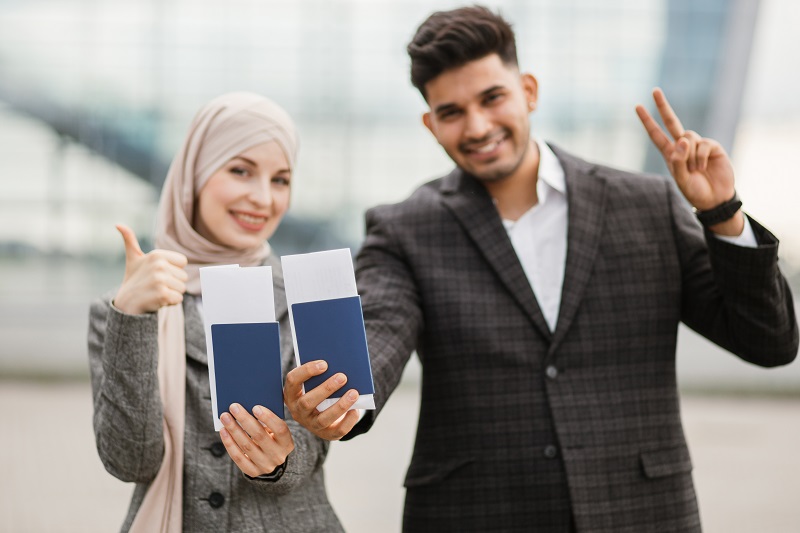 Smiling Muslim woman in hijab and Arab man, posing on camera outside modern airport terminal, demonstrating their passports and tickets while waiting flight. Ready for trip. Business people
